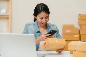 Mujer revisando un paquete con el móvil junto a un portátil y cajas de envío